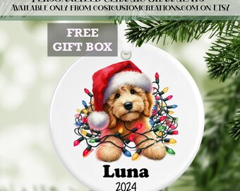 May include: A white ceramic Christmas bauble featuring a cartoon illustration of a dog wearing a Santa hat and tangled in Christmas lights. The dog's name, "Luna", and the year "2024" are printed below the dog.