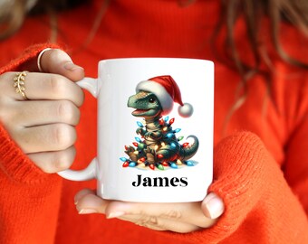 May include: White ceramic mug featuring a cartoon dinosaur wearing a Santa hat and wrapped in Christmas lights. The name "James" is printed in black on the mug. The mug is held by a person wearing an orange sweater.