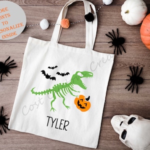 Puede incluir: Bolsa de tela blanca con un esqueleto de dinosaurio verde que sostiene una calabaza tallada. El dinosaurio camina hacia la derecha y hay tres murciélagos negros volando sobre él. El texto "TYLER" está impreso debajo del dinosaurio.