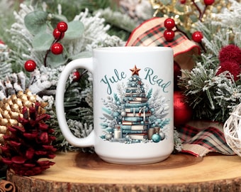 May include: White ceramic mug with a Christmas tree design made of books, with the words "Yule Read" in teal script. The mug is decorated with a star, ornaments, and winter foliage. The mug is on a wooden surface with Christmas decorations.