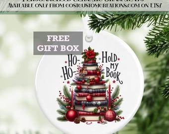 May include: A white ceramic Christmas ornament featuring a festive design of a Christmas tree made of books, red berries, and baubles. The ornament has the text "Hold my Book" and "Ho Ho Ho". The text "Free Gift Box" is also visible.