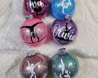 Op de afbeelding: Zes glinsterende kerstballen met gepersonaliseerde namen en een silhouet van een turnster in handstand. De ballen zijn roze, paars, blauw, turquoise en zilver.