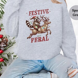 Op de afbeelding: Een grijs sweatshirt met een feestelijke wasbeer die op een rendier met kerstverlichting rijdt. De tekst op het sweatshirt luidt "Festive But Feral".