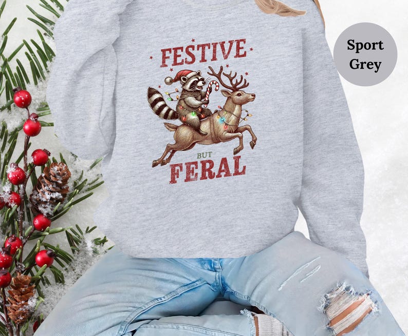 Op de afbeelding: Een grijs sweatshirt met een feestelijke wasbeer die op een rendier met kerstverlichting rijdt. De tekst op het sweatshirt luidt "Festive But Feral".