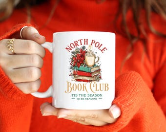 May include: White ceramic mug featuring the text "NORTH POLE BOOK CLUB" with an illustration of books, poinsettias, and a cup of whipped cream. The mug also has the text "TIS THE SEASON TO BE READING".