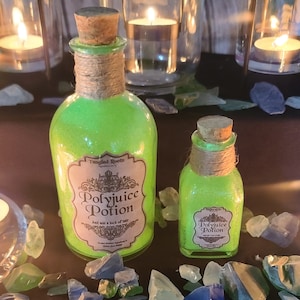 Puede incluir: Dos botellas de vidrio verde con tapones de corcho y cuerda envuelta alrededor de los cuellos. Las botellas están llenas de un líquido verde y tienen etiquetas que dicen "Polyjuice Potion" y "Just add a lock of hair". Las botellas están sobre una superficie con fragmentos de vidrio verde y azul.