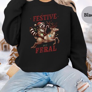Op de afbeelding: Zwart sweatshirt met een feestelijk maar wild ontwerp. Een wasbeer met een kerstmanmuts rijdt op een rendier met kerstverlichting. De tekst luidt "Festive But Feral".