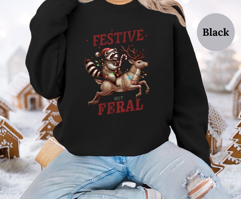 Op de afbeelding: Zwart sweatshirt met een feestelijk maar wild ontwerp. Een wasbeer met een kerstmanmuts rijdt op een rendier met kerstverlichting. De tekst luidt "Festive But Feral".
