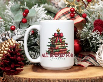 May include: White ceramic mug with a Christmas tree design made of books, a red bow, and a gold star. The text "ALL BOOKED FOR Christmas" is printed below the tree. The mug is on a wooden surface with festive decorations.