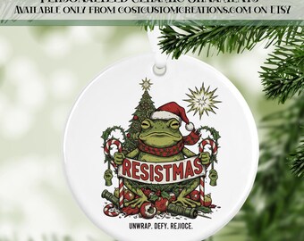 May include: White ceramic Christmas ornament featuring a frog wearing a Santa hat and scarf, holding a banner that reads "RESISTMAS." The ornament includes a Christmas tree, candy canes, and the text "UNWRAP. DEFY. REJIOCE."