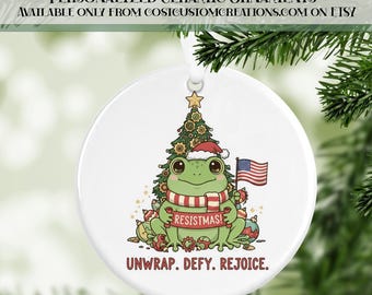 May include: A white ceramic Christmas ornament featuring a cartoon frog wearing a Santa hat and holding a US flag. The frog is in front of a Christmas tree. The text "RESISTMAS!" is on a scarf. The ornament reads "UNWRAP. DEFY. REJOICE."