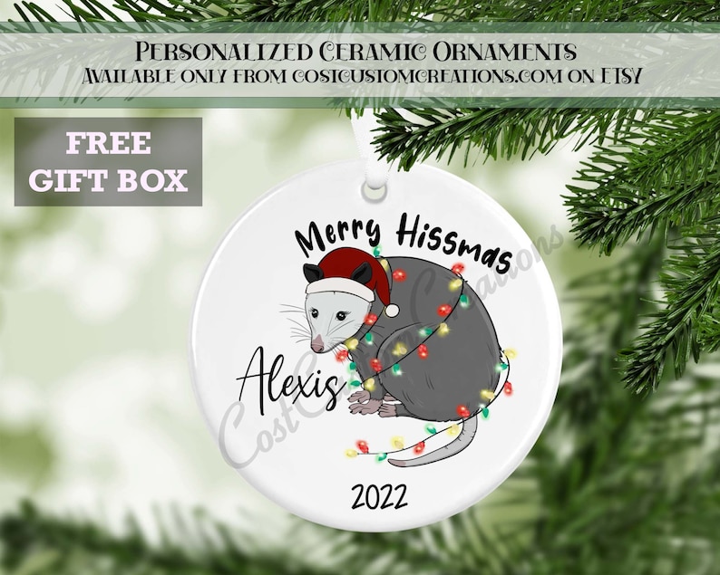 May include: A white ceramic Christmas bauble featuring a grey opossum wearing a red Santa hat and surrounded by colourful Christmas lights. The bauble reads "Merry Hissmas" and "Alexis" in black text. The year "2022" is printed in black at the bottom of the bauble.