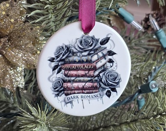 May include: White ceramic Christmas ornament with a stack of books and gray roses. The books have the text "STFUATTDLAGG" and "DARK ROMANCE" on a banner. A magenta ribbon is attached for hanging.