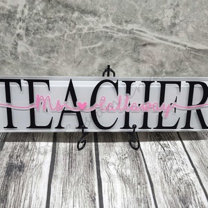 Peut inclure: Une plaque en acrylique blanche avec le mot "TEACHER" en lettres noires. Le mot "Ms." est écrit en cursive rose avec un petit cœur rose au-dessus. La plaque est sur un support noir.
