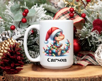 May include: White ceramic mug featuring a cartoon dinosaur wearing a Santa hat and Christmas lights. The dinosaur is sitting with the name "Carson" printed in black below. The mug is set against a festive holiday backdrop.