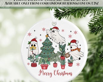 May include: White ceramic Christmas ornament with a festive design. Features three ghost characters wearing Santa hats, a decorated Christmas tree, and potted plants. The words "Merry Christmas" are written in red.