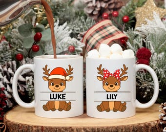 May include: Two white ceramic mugs with reindeer designs. One mug has a reindeer wearing a Santa hat and the name "LUKE". The other mug has a reindeer with a red polka dot bow and the name "LILY". One mug is filled with marshmallows.