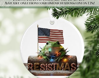 May include: A round ceramic ornament featuring a green frog with string lights, an American flag, and the word "RESISTMAS" in candy cane letters. The ornament has a white background and a silver hanging loop.