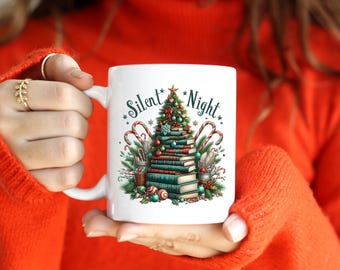 May include: White ceramic mug featuring a Christmas tree design made of books, candy canes, and ornaments. The mug has the words "Silent Night" printed on it. The mug is held by a person wearing an orange sweater.