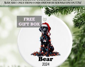 May include: A white ceramic Christmas ornament featuring a black Great Dane dog wearing a Santa hat and tangled in Christmas lights. The dog's name, "Bear", and the year "2024" are written below the dog.  "FREE GIFT BOX" is written in a pink box at the top of the ornament.