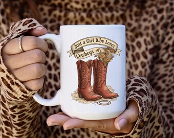 May include: White ceramic mug featuring a design with a pair of red cowboy boots, a bouquet of flowers, and the text "Just a Girl Who Loves Cowboys" in a banner. The mug is held in a person's hands.