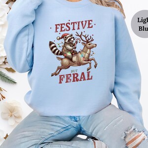 Op de afbeelding: Lichtblauwe trui met een feestelijke wasbeer die op een rendier met kerstverlichting rijdt. De tekst op de trui luidt "Festive But Feral".