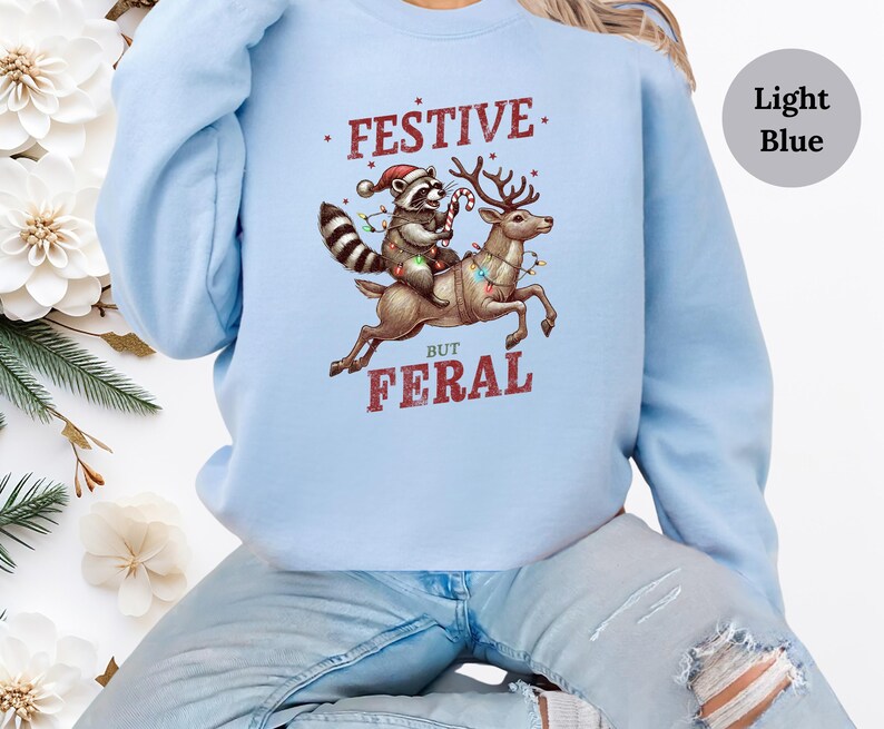 Op de afbeelding: Lichtblauwe trui met een feestelijke wasbeer die op een rendier met kerstverlichting rijdt. De tekst op de trui luidt "Festive But Feral".