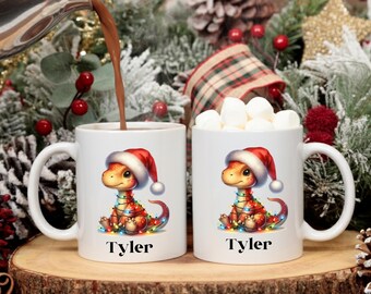 May include: Two white ceramic mugs with a Christmas dinosaur design. Each mug features a cartoon dinosaur wearing a Santa hat and wrapped in Christmas lights. The name "Tyler" is printed on each mug.
