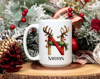 May include: White ceramic mug with a festive Christmas design. The mug features a brown letter "N" decorated with reindeer antlers, a red and green scarf, and the name "NATHAN" in black text. Christmas-themed decorations are in the background.