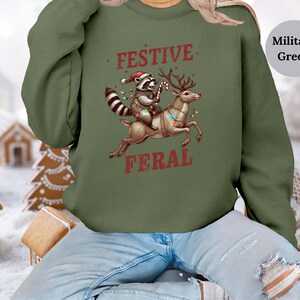 Op de afbeelding: Een groen kersttrui met een wasbeer die een kerstmanmuts draagt ​​en op een rendier rijdt met de tekst "Festive Feral" erop gedrukt.