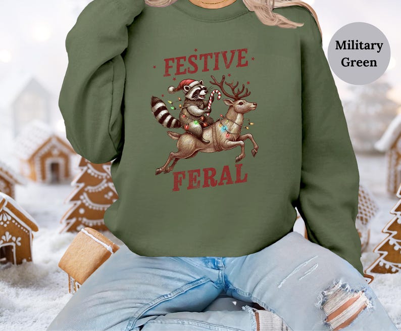 Op de afbeelding: Een groen kersttrui met een wasbeer die een kerstmanmuts draagt ​​en op een rendier rijdt met de tekst "Festive Feral" erop gedrukt.