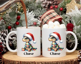 May include: Two white ceramic mugs with a Christmas dinosaur design. Each mug features a cartoon dinosaur wearing a Santa hat and Christmas lights. The name "Chase" is printed on the bottom of each mug. One mug is being filled with a hot beverage, the other is filled with marshmallows.