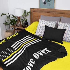 Puede incluir: Manta de tiro negra y amarilla con una fina franja amarilla sobre el diseño de la bandera estadounidense. El texto "Slow Down Move Over" está impreso en blanco sobre la manta.