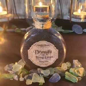 May include: A dark purple glass bottle with a cork stopper and a label that reads "Draught of Living Death - Sleeping Potion - Gives the appearance of death - Trusted Premium Ingredients & Elixir since 1872" with a skull and crossbones design. The bottle is surrounded by small pieces of sea glass.