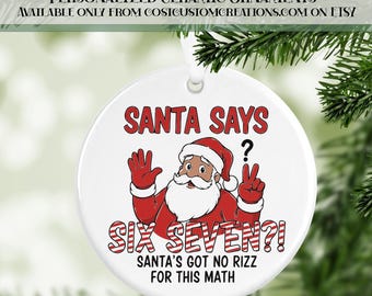 May include: A white ceramic Christmas ornament with a Santa Claus graphic. The text "SANTA SAYS? SIX SEVEN?! SANTA'S GOT NO RIZZ FOR THIS MATH" is displayed in red and white. The ornament is round and has a silver string for hanging.