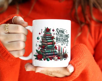 May include: White ceramic mug featuring a Christmas tree made of stacked books, adorned with ornaments and greenery. The mug has the text "Spine-Chilling Holidays" and is held by a person wearing an orange sweater.