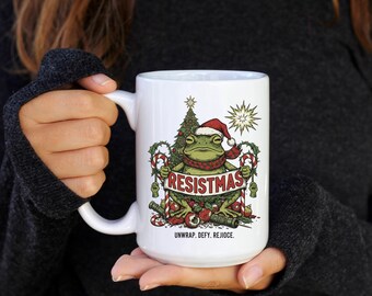 May include: White ceramic mug featuring a frog wearing a Santa hat and scarf, holding a banner that reads "RESISTMAS." The mug has a Christmas tree and star design. The text "UNWRAP. DEFY. REJOICE." is printed below the frog.