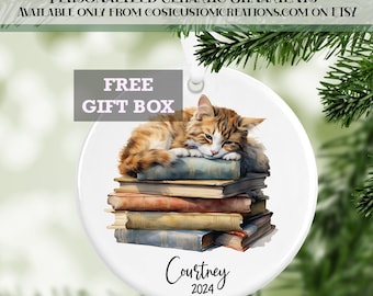 May include: White ceramic Christmas ornament featuring a watercolor illustration of a ginger cat sleeping on a stack of vintage books. The ornament is personalized with the name "Courtney" and the year "2024".