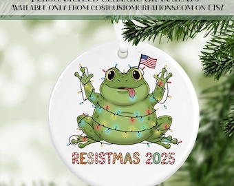 May include: White ceramic ornament featuring a cartoon frog decorated with Christmas lights and an American flag. The frog has its tongue out and the text "RESISTMAS 2025" is at the bottom. The top text reads "PERSONALIZED CERAMIC ORNAMENTS".