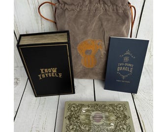 May include: An oracle card set with a black box that reads "KNOW THYSELF" in gold lettering, a deck of cards, a book titled "The Two-Penny Oracle", and a brown velvet drawstring bag with an elephant design.