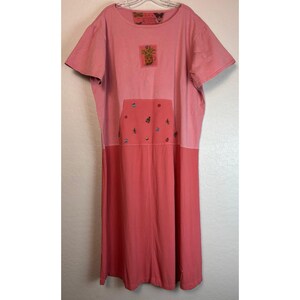 May include: A two-tone pink dress with short sleeves. The top section is a lighter pink, while the bottom is a darker shade. A square patch with a floral design is sewn on the chest. A pocket with butterfly and dot embellishments is on the front.