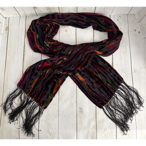 May include: A black scarf with a velvet zebra stripe pattern in shades of red, orange, and green. The scarf has long black fringe at both ends. The scarf is made of a soft, flowing material.