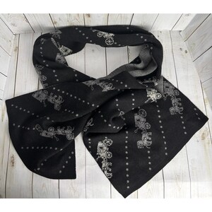 May include: Black scarf with a repeating pattern of horse-drawn carriages in gray. The scarf has a dotted design and is draped on a light wood surface. The scarf is made of a soft material.