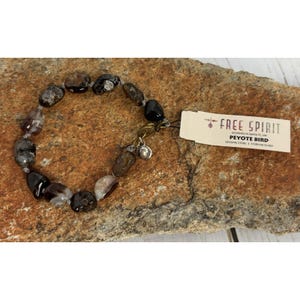 Op de afbeelding: Een kralenarmband met donkerbruine, zwarte en heldere stenen. De armband heeft een zilveren sluiting en bedel. Een label leest "FREE SPIRIT PEYOTE BIRD" en "GENUINE STONE | STERLING SILVER."
