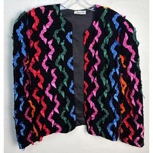 May include: A black open-front jacket featuring vertical rows of ruffled ribbon trim in vibrant shades of pink, blue, green, red, and orange. The garment has long sleeves, a solid black interior lining, and a label reading Miss H at the neckline.