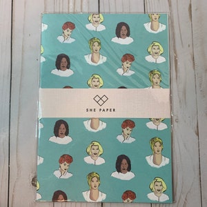 May include: A stack of paper featuring a teal background with repeating illustrations of women's faces. The paper is wrapped with a band that reads "SHE PAPER" in white text.