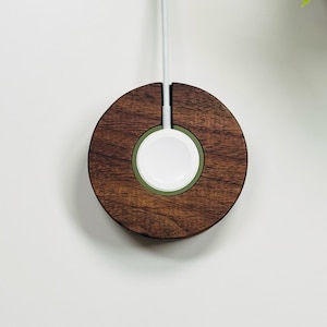 May include: A round, wooden Apple Watch charging dock. The dock is made of dark brown wood with a white charging pad and a green accent ring. A white charging cable extends upwards from the dock.
