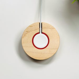 May include: A wooden Apple Watch charging stand. The stand is circular with a light wood finish and a red accent ring around the white charging surface. A gray charging cable extends from the top.
