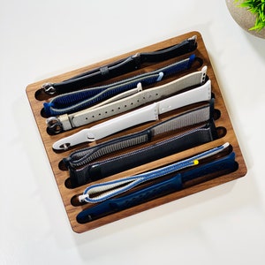 May include: A wooden watch band organizer with six slots. The slots are filled with different colored watch bands, including black, blue, gray, and white.