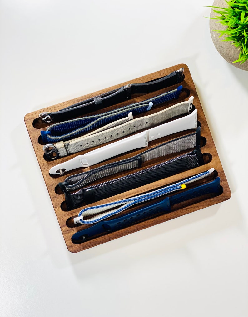 May include: A wooden watch band organizer with six slots. The slots are filled with different colored watch bands, including black, blue, gray, and white.